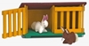 Cage à lapins jouet de ferme rouge, verte et jaune avec une des 2 portes ouvertes, un petit lapin blanc en laine dans la cage, un lapin brun devant la cage.