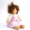Petite poupée de 25 cm, assise, ayant des cheveux de mohair brun et des yeux bruns, portant un t-shirt blanc à manches longues et une robe chasuble rose.
