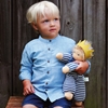 Un petit garçon blond à chemise bleu clair et short bleu foncé est assis sur un banc. Il tient dans le bras gauche une poupée en tissu éponge rayé blanc et bleu foncé, avec des cheveux blonds en mohair et une couronne en feutre de laine jaune.