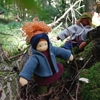 Standing outside at the foot of a tree, a Nanchen doll with blue eyes, red hair with navy blue headband, burgundy dress, green pants and petrol blue wool fleece hooded jacket.